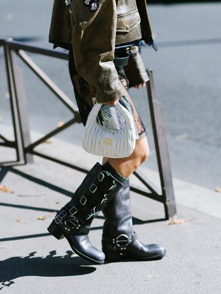 A woman wear a brown leather jacket, white miu miu bag, black engineer boots, and a skirt.