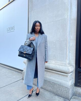 Woman wearing gray coat, blue jeans, black slingback heels, and a black Chanel bag stands against wall.