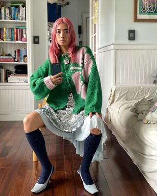 Woman sitting on chair in bedroom wearing green athletic jacket, sequin dress, knee-high navy socks and white slingback heels.