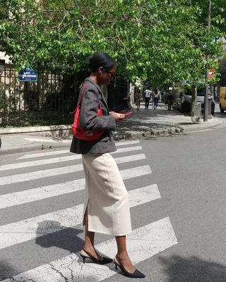 Woman wearing white maxi skirt, grey suit jacket, red handbag and black slingback heels standing on crosswalk.