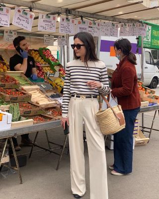 French woman at an outdoor market wearing a striped sweater, cream trousers, and a black belt carrying a raffia bag