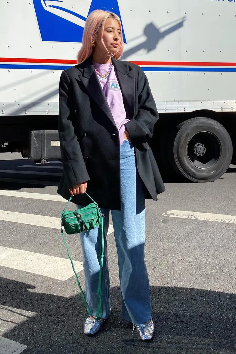 Pessoa vestindo blazer oversized preto, camiseta roxa, calça jeans ampla e sapatos prateados. Acessório: bolsa verde. Tendências de moda urbana com mistura de peças clássicas e modernas, realçando um estilo descontraído e criativo.