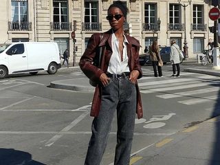 Woman wearing jeans, a white button down, and a brown leather coat.