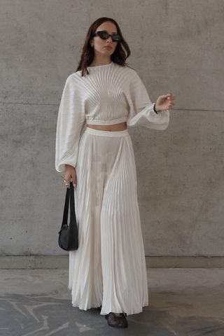 a woman wearing a white pleated maxi skirt and matching pleated top and black shoulder bag and black flats
