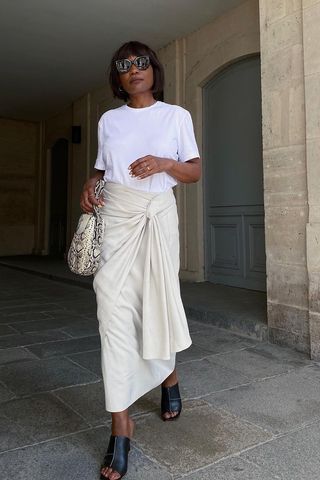 a photo of a woman wearing a white wrap skirt with a white t-shirt and black mules and a gray and white snakeskin bag