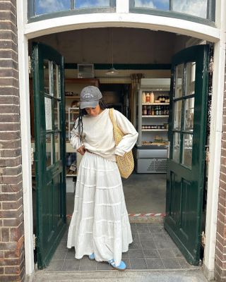 style influencer stands in a grocery story doorway wearing a spring outfit with a baseball cap, sweatshirt, white tiered maxi skirt, and blue Adidas Gazelle sneakers