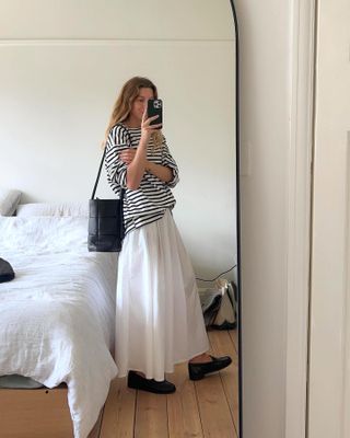 British fashion influencer taking a mirror selfie with a striped T-shirt, white maxi skirt, woven bucket bag, and loafers