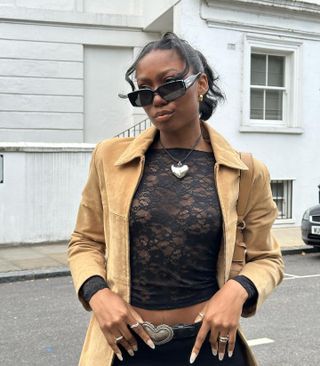 A woman posing on the street in front of a white building and wearing black sunglasses, a black lace top, a tan suede jacket, and silver heart necklace and belt
