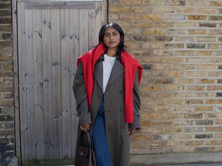 Woman wearing a long coat with jeans, a white tee, and a red sweater draped over her shoulders.