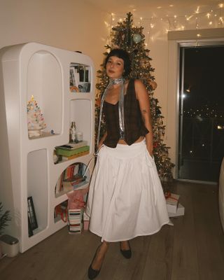 Alyssa wears a brown vest, silver neck tie, a white skirt, and ballet flats.