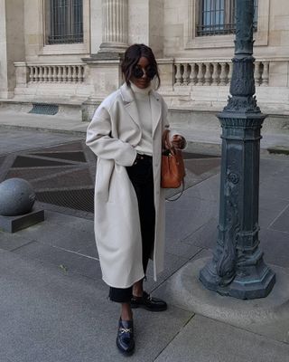 A woman wears a gray wool coat, white turtleneck, black cropped pants, and black loafers.