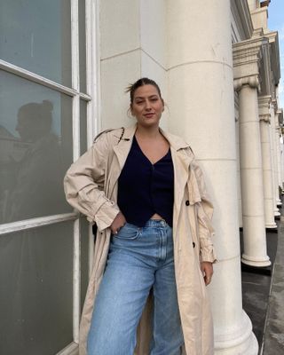 A woman wears a black tailored vest, beige trench coat, and baggy blue jeans.