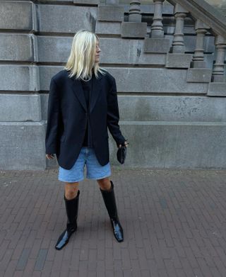 A woman wears an oversize black blazer, denim jorts, and black knee-high boots.