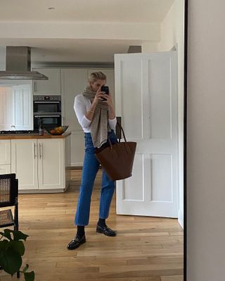 A woman wears a beige scarf, white long-sleeve shirt, blue jeans, black socks, a brown suede tote bag, and black loafers.