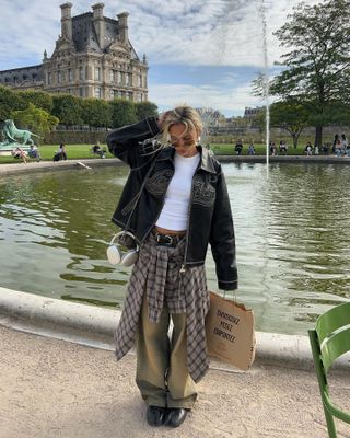 A woman wears a brown leather distressed jacket, white t-shirt, plaid shirt over khaki pants, with black tabi shoes.