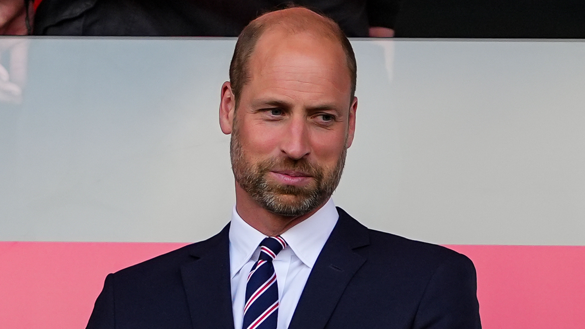Prince William wearing a suit and tie in front of a pink and white background