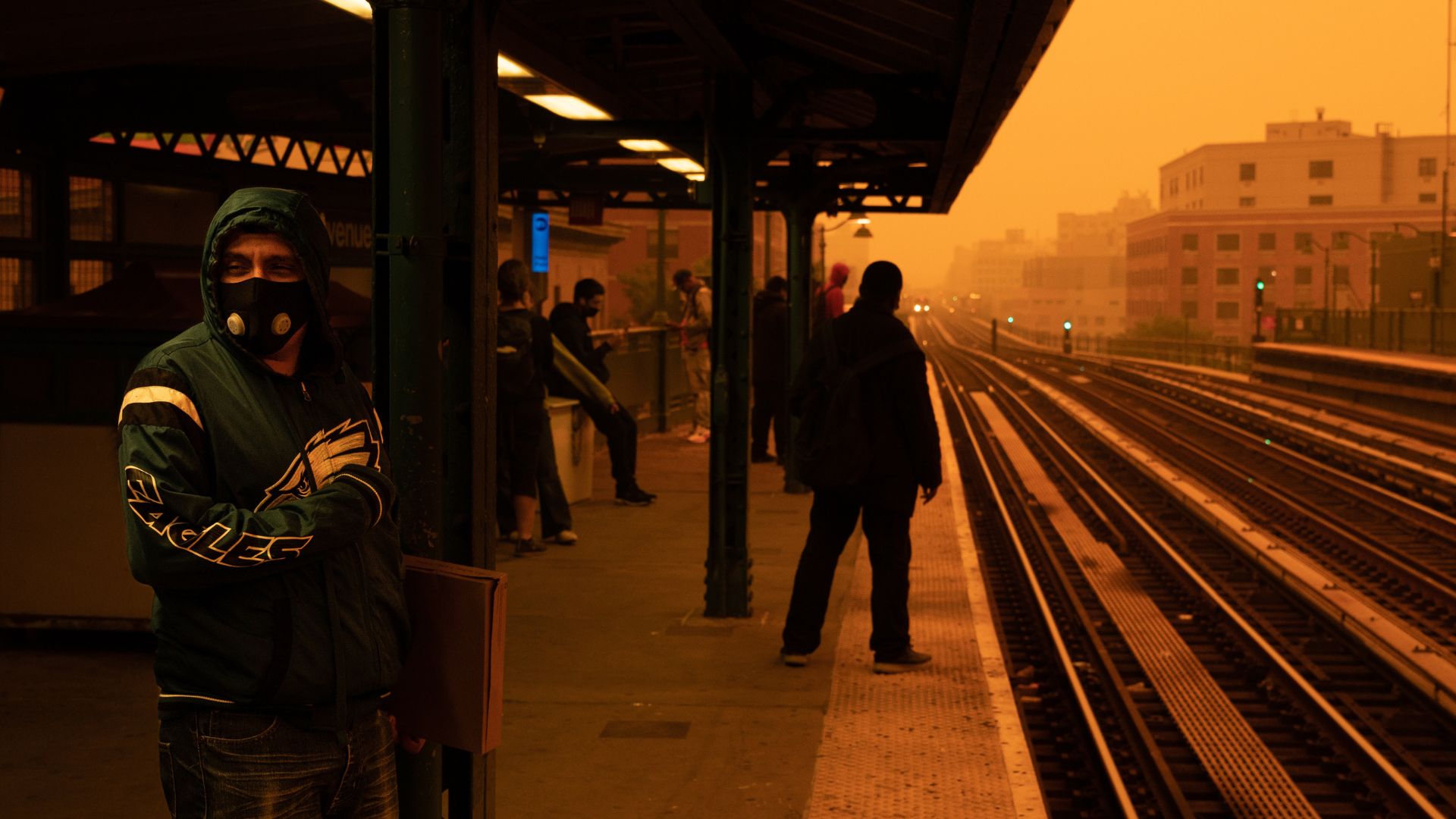 photo shows people waiting at an above ground subway station in the bronx. A man in the foreground wears a heavy duty respirator, and the sky and air surrounding the station appears dark orange