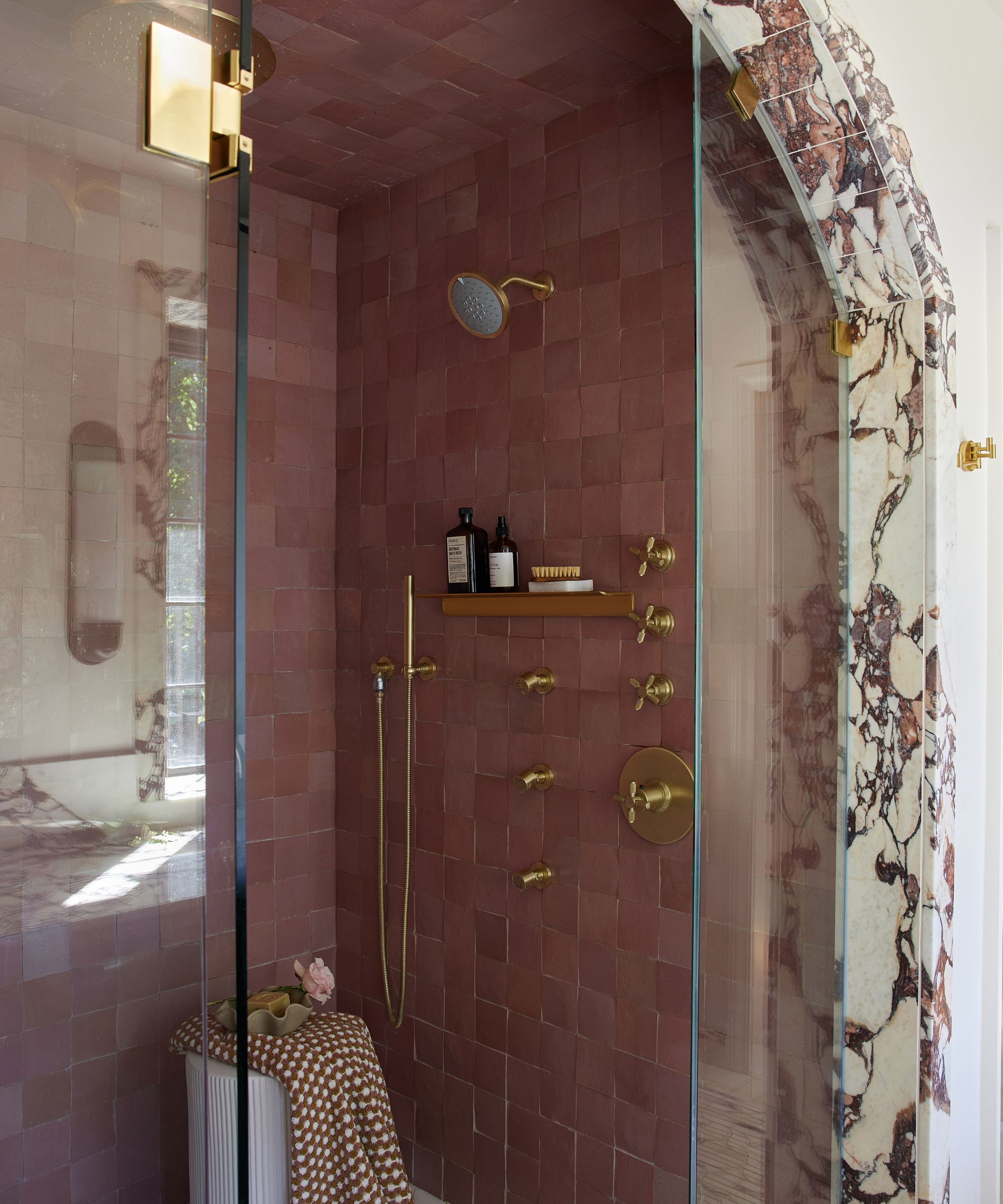 A shower with pink zellige tiles, brass fixtures, and a marble arch at the entrance.