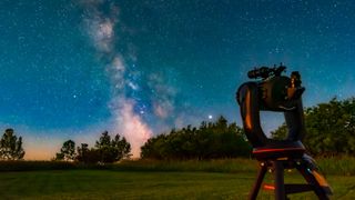 A telescope on a tripod sits in a grassy field looking at the pink glow of the Milky Way galaxy past some trees with a light blue night sky behind