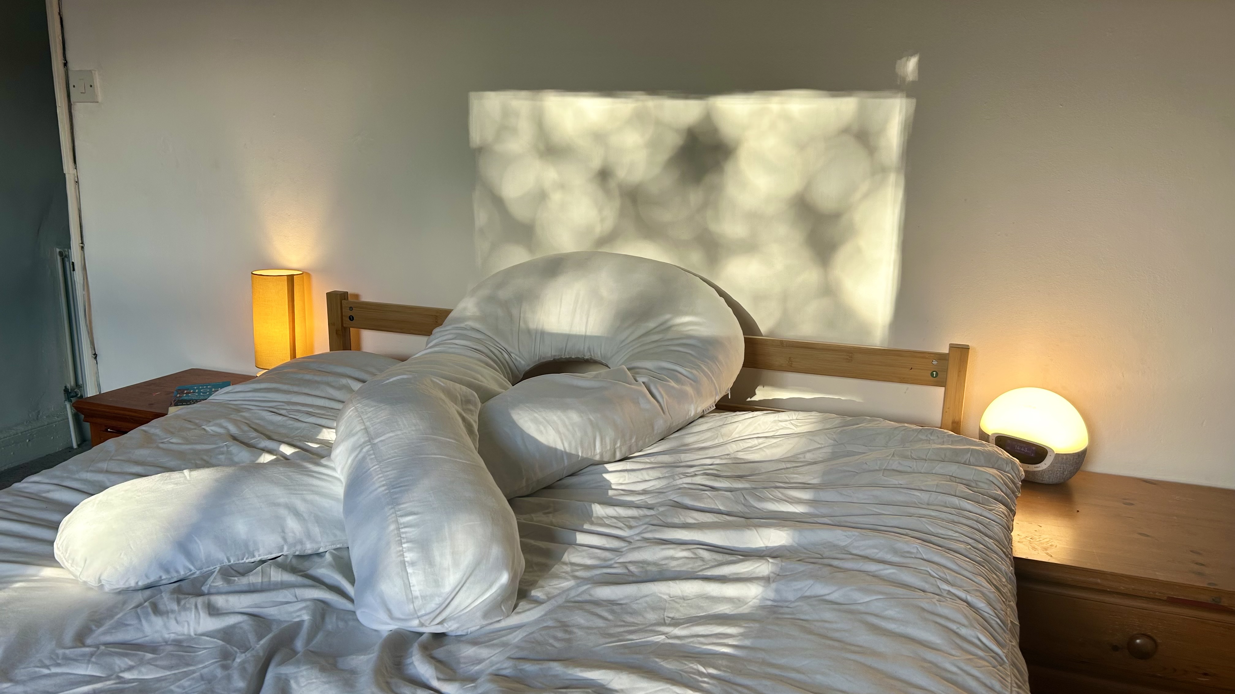 The Yana 360 Body Pillow crossed over itself on top of a bed dressed in white bedding
