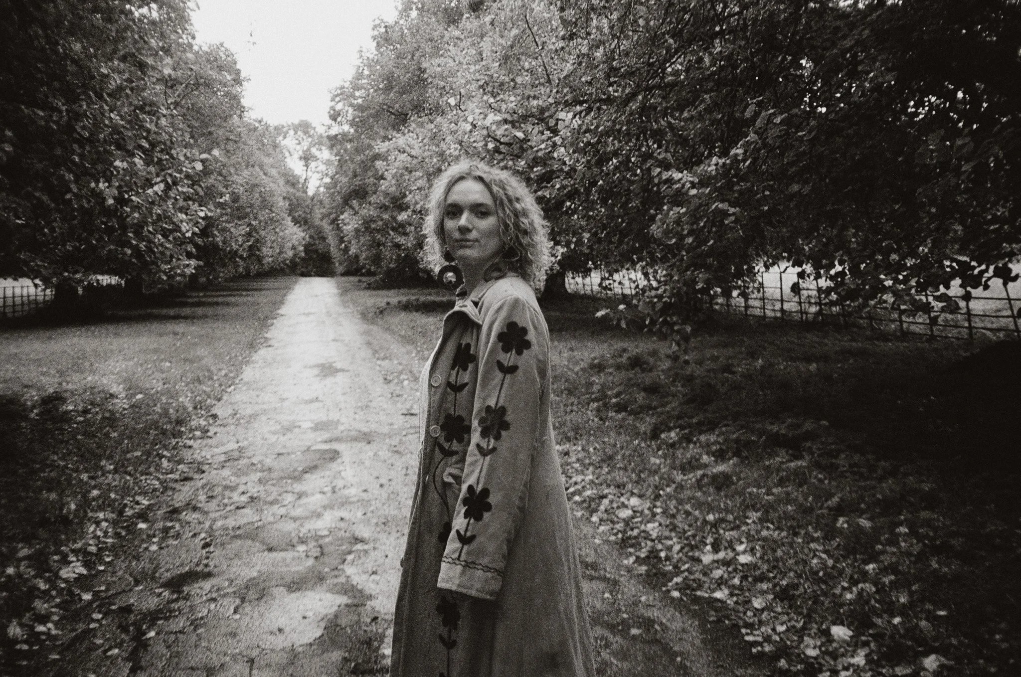 A black-and-white portrait of a young woman with curly blonde hair standing in a park sideways while wearing a flower-pattern suede coat.