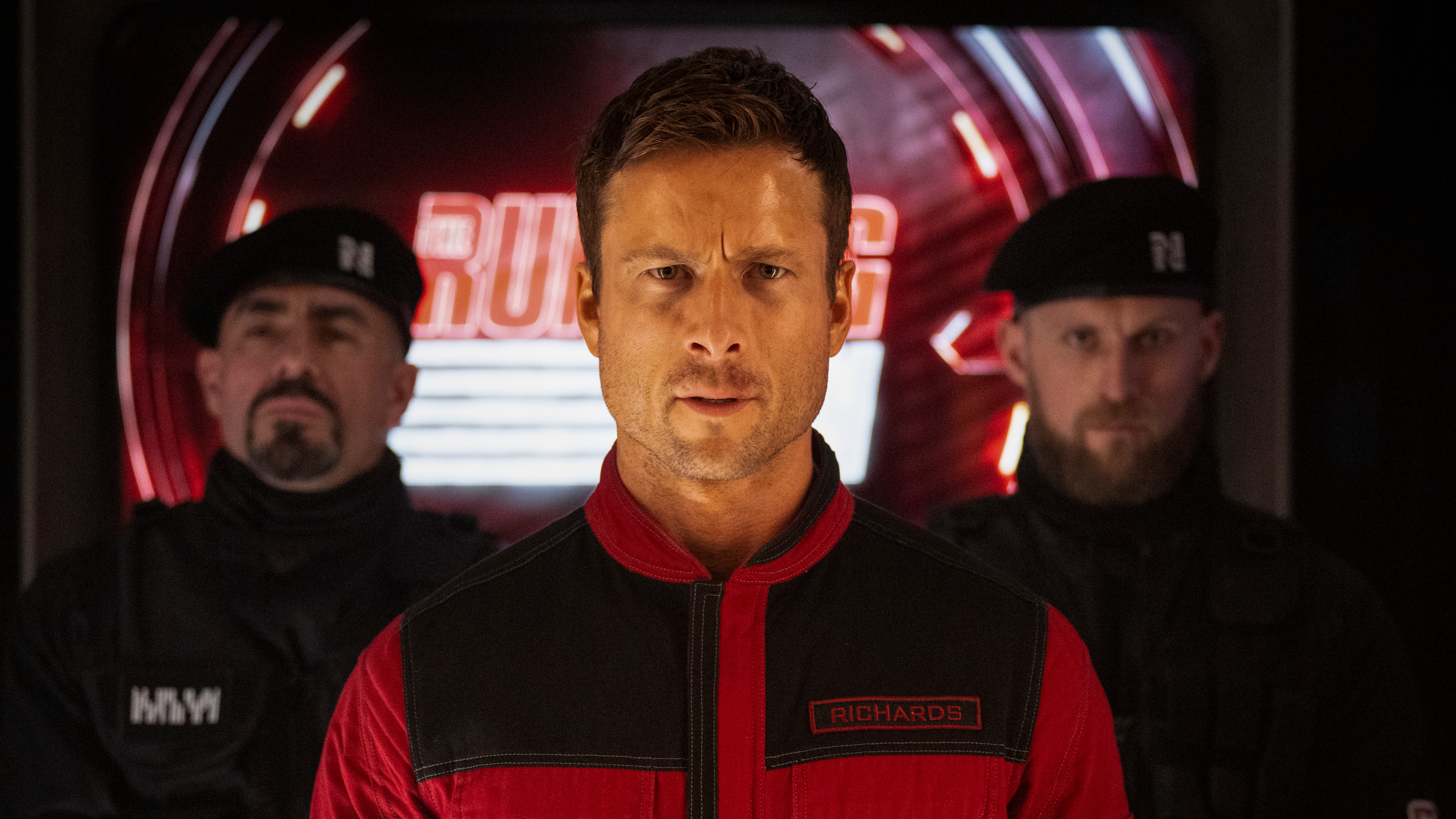 Ben Richards (Glen Powell) in his red jumpsuit flanked by guards in a scene from "The Running Man" (2025)