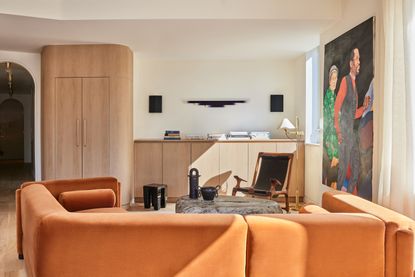 A living room with a large orange sofa, light wooden built in cabinetry, and a shelf decorated with magazines