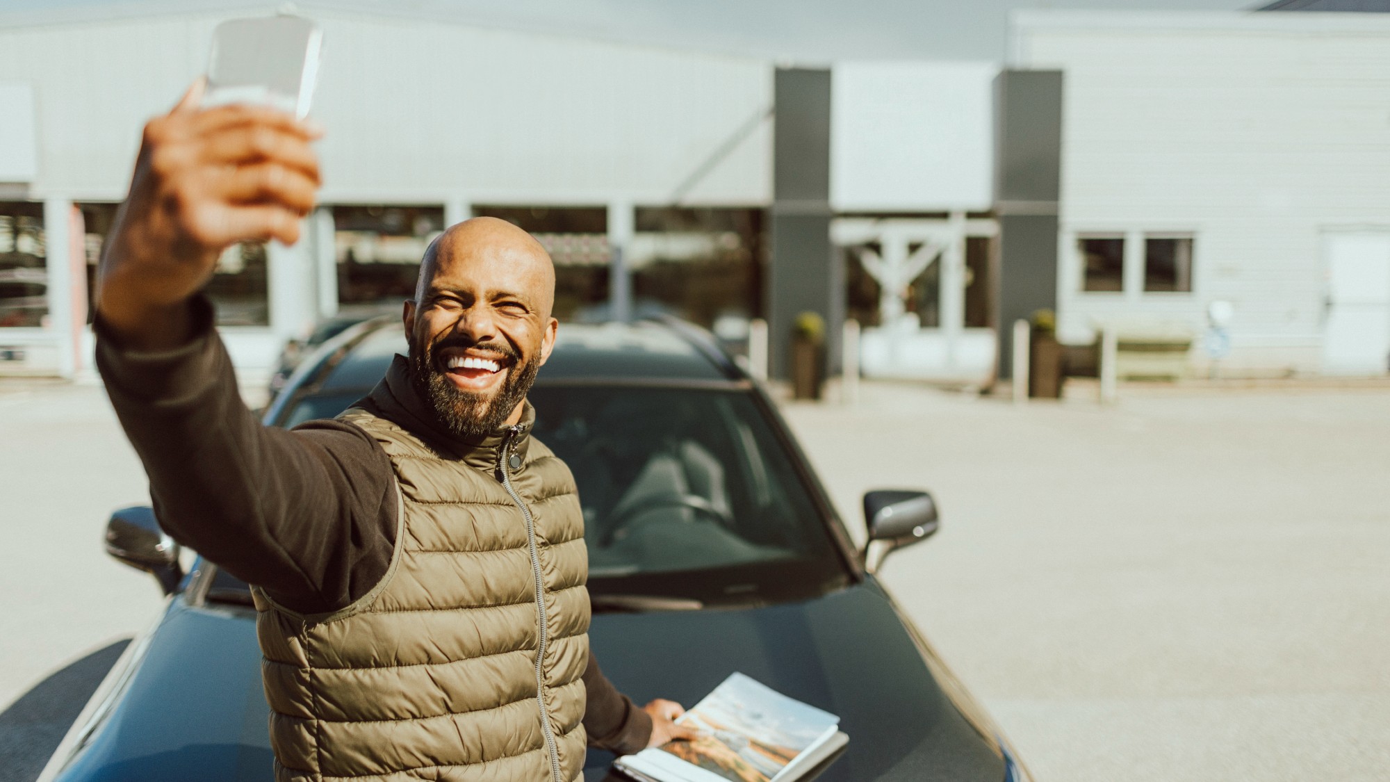 Cheerful man taking a selfie with his new car outside of a showroom