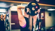 Woman exercising using weight plate in the gym. She holds the weight plate behind her head.