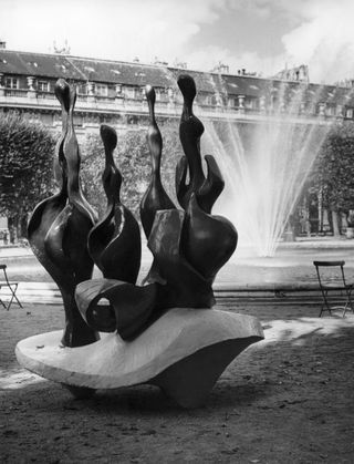 In a park, an organically shaped, abstract sculptures features multiple anthropomorphic structures on a wavy pedestal. Behind them is a sprouting fountain.