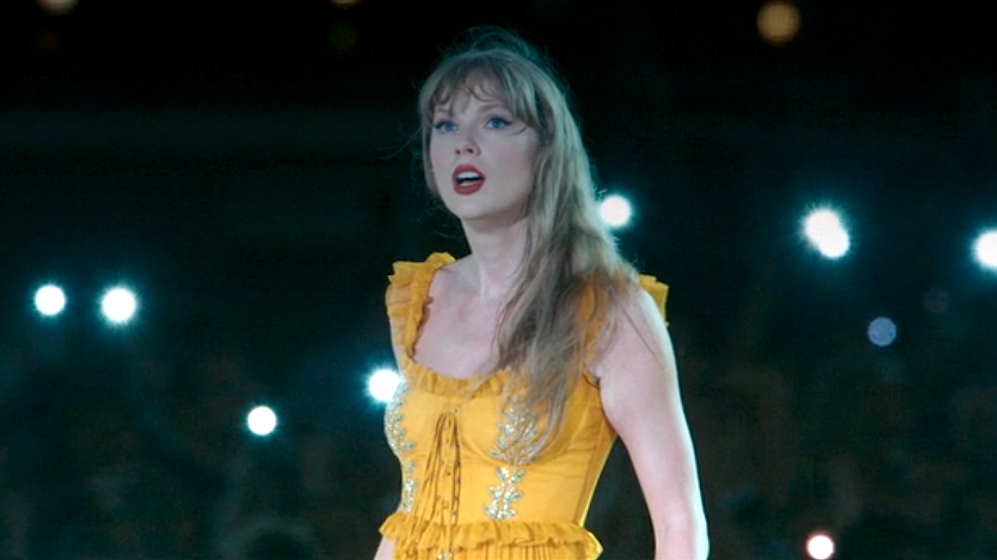 Taylor Swift singing "Marjorie" in a yellow dress with cell phone lights shining in the background