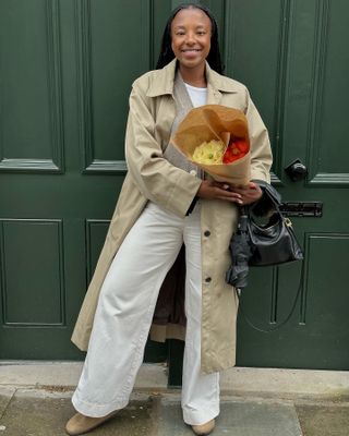 fashion influencer Taffy Msipa poses in front of a green door with flowers wearing a trench coat, neutral cardigan, white jeans, and tan suede clogs