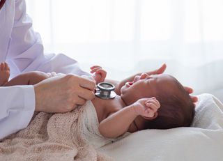 A medical professional holding a stethoscope to the chest of an infant lying on its back. 