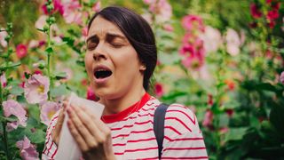 Woman in red and white striped shirt sneezes into a tissue while standing among flowers.