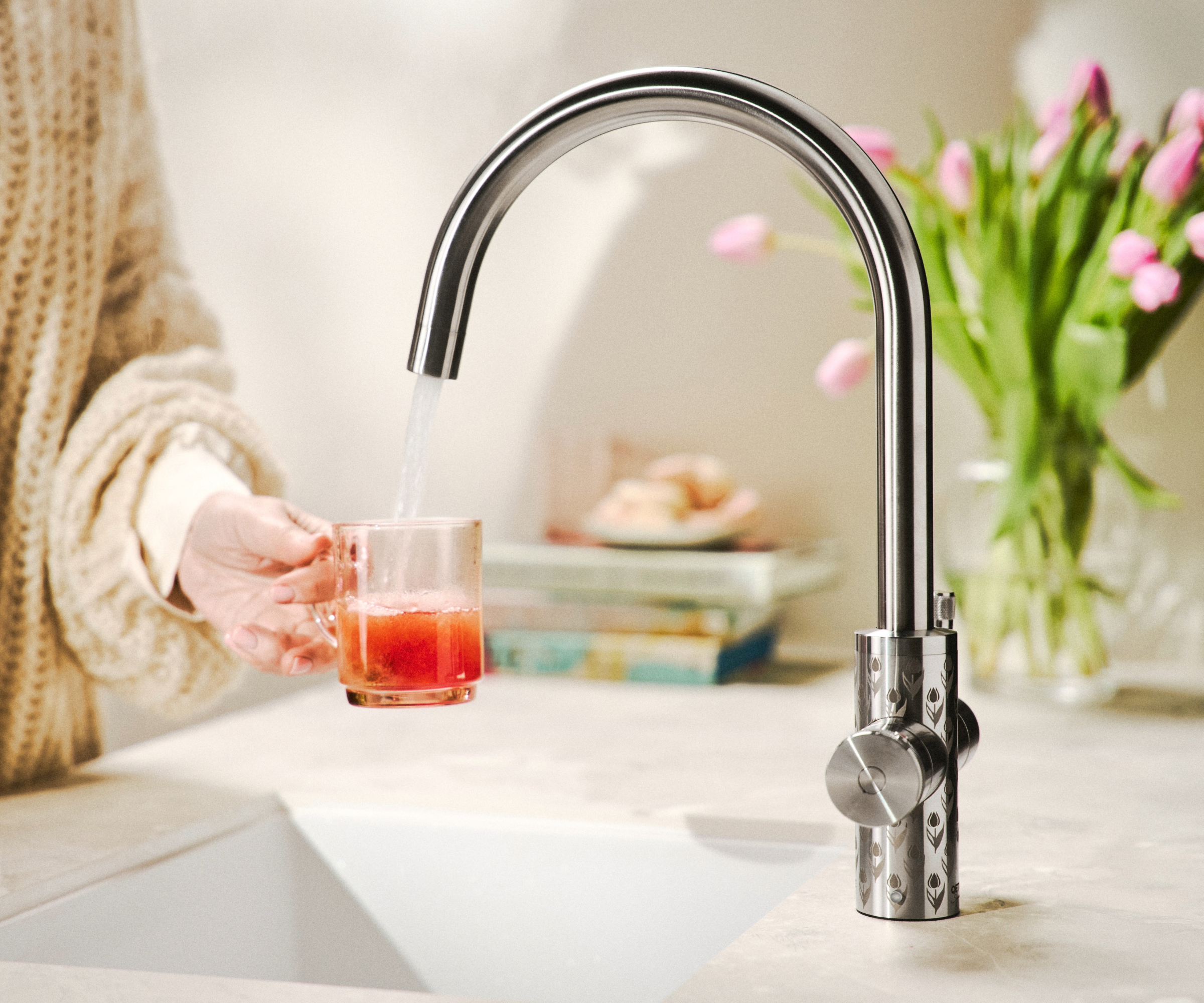 Person holding a cup under the QETTLE Signature Pattern Tulips 4 in 1 100C Boiling Water Tap with flowers in background
