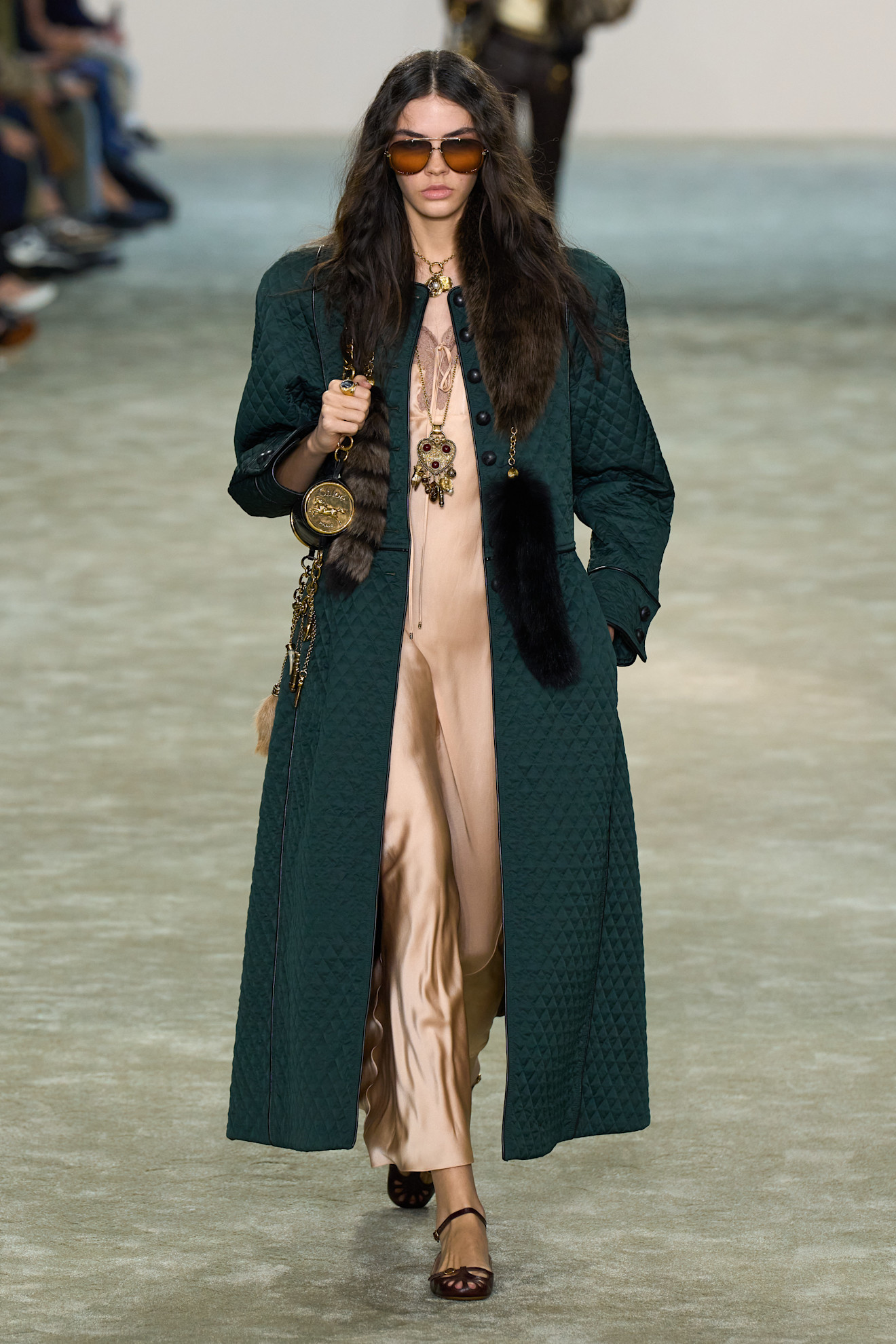 Fur accessory trend shown in Chloe&#039;s fall-winter 2025 collection in a photo of a model walking down the runway wearing an emerald green coat over a nude maxi slip dress styled with red flats, a hobo shoulder bag, aviator sunglasses, layered gold necklaces, and a fox tail scarf
