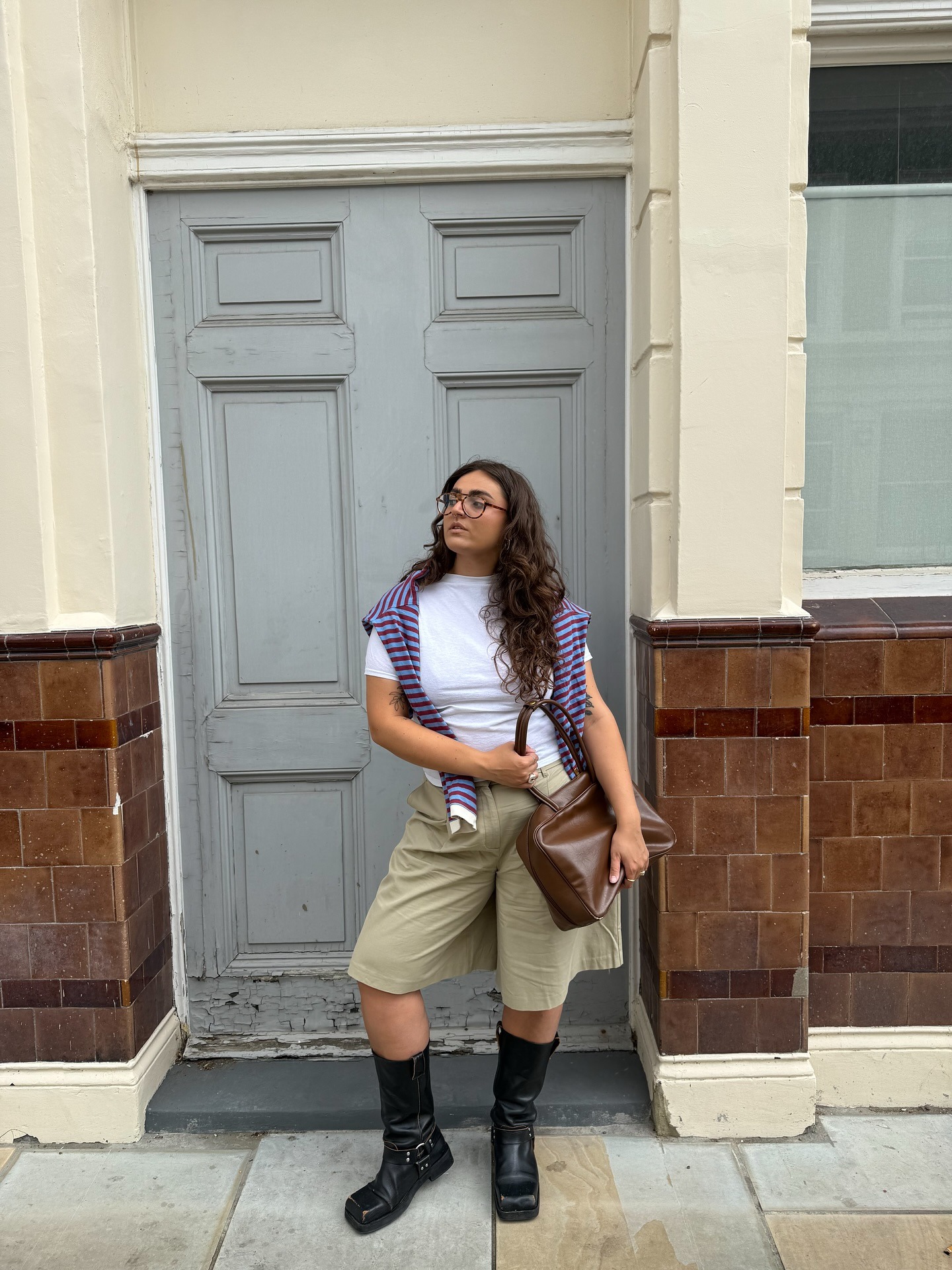 Fashion person Izabella wears khaki Bermuda shorts, black leather biker boots a white t-shirt a stripped jumper thrown over the shoulder and a warm-brown square bag in a recent image from her Instagram.