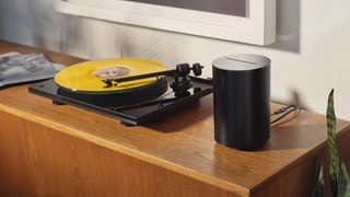 Der Sonos Era 100 in Schwarz auf einem Holzregal neben einem gelben Plattenspieler