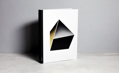 Hardcover book with a black geomtric designed shape object drawn on the face against a white background . Photographed on a grey surface , against a grey wall