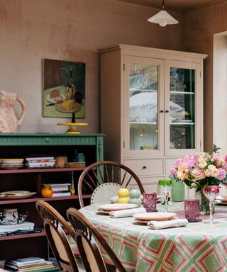 A dining area with terracotta walls, a green bookshelf with colorful books, a beige cabinet, and a dining table with a green and pink patterned cloth with plates and napkins on and dark wooden chairs