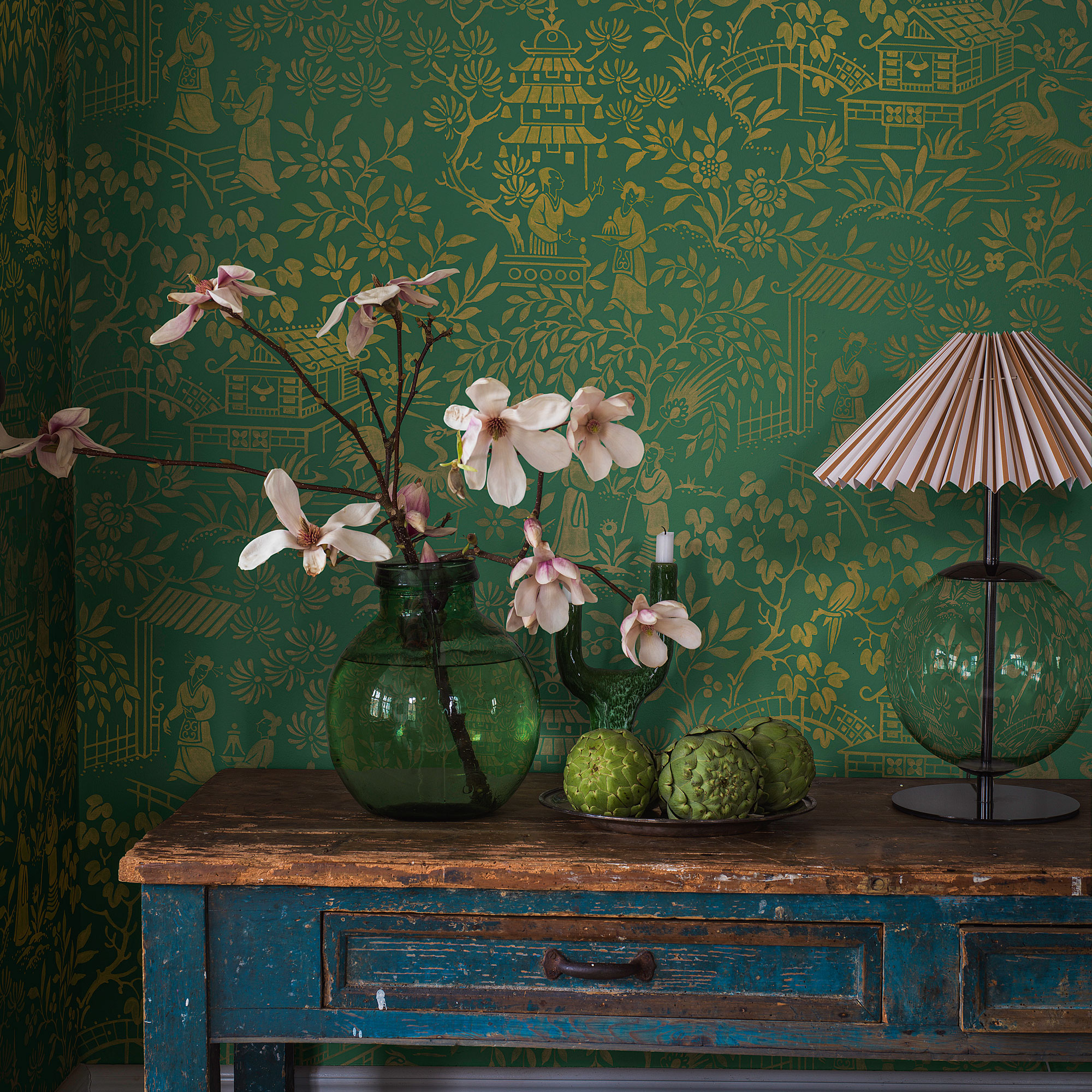 Green and gold wallpaper with a blue antique side table, green case with magnolia branches and a clear lamp with pink shade.