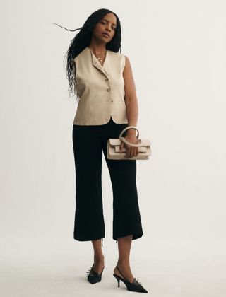 Model and content creator Maya Moore, who is missing her right arm, in a campaign for Anthropologie's adaptive line wearing a tailored tan vest with crop flares, a clutch purse, and kitten heel slingback heels.