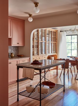 Shot of an open-concept kitchen that leads into a casual dining room. The cabinets are painted in a dusty pink and there is a small, open kitchen island in the center of the room.