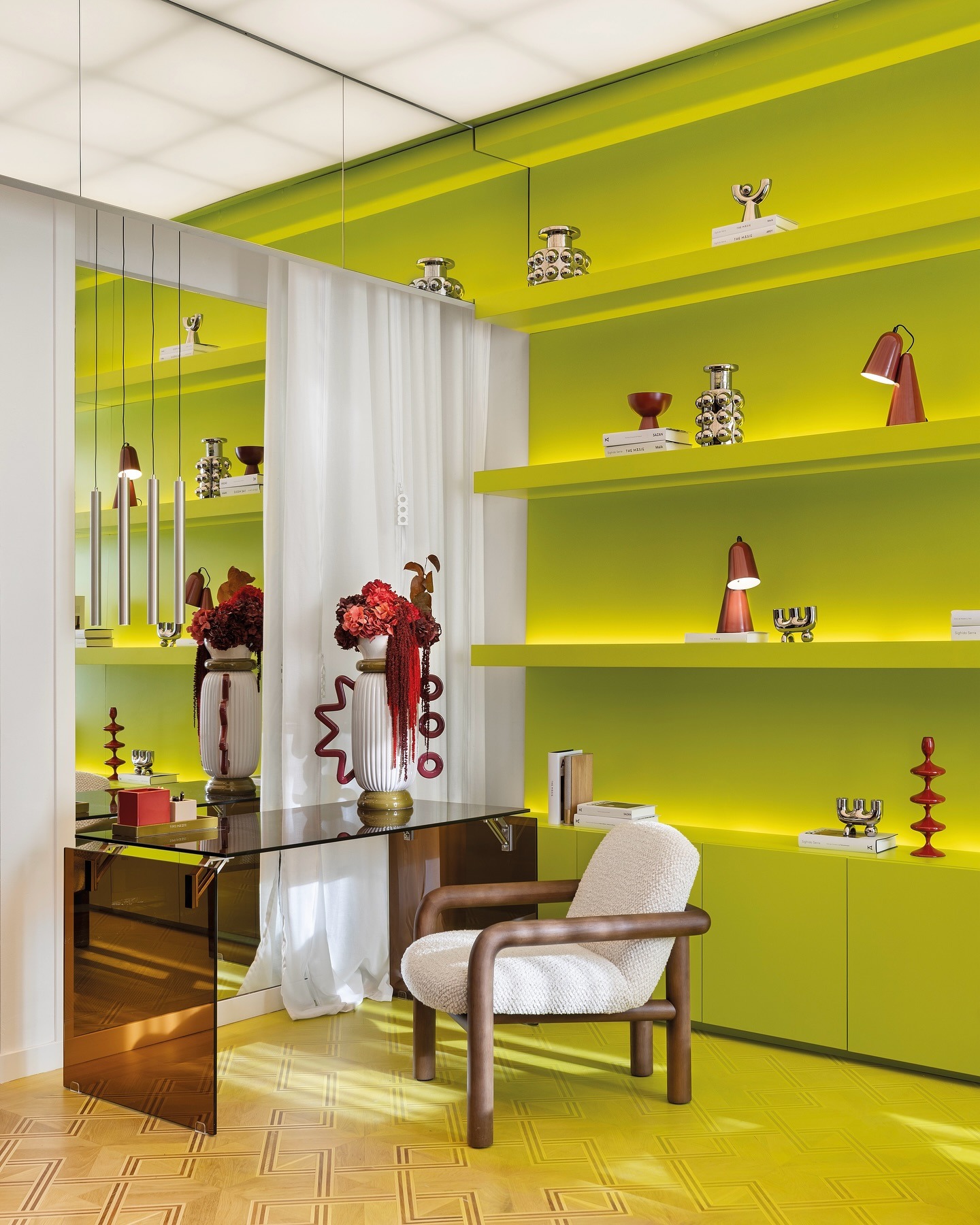 Image of an office room with lime green built-in shelving covering the walls and a brown glass desk with a white wooden desk chair. There is a white curtain behind the desk.