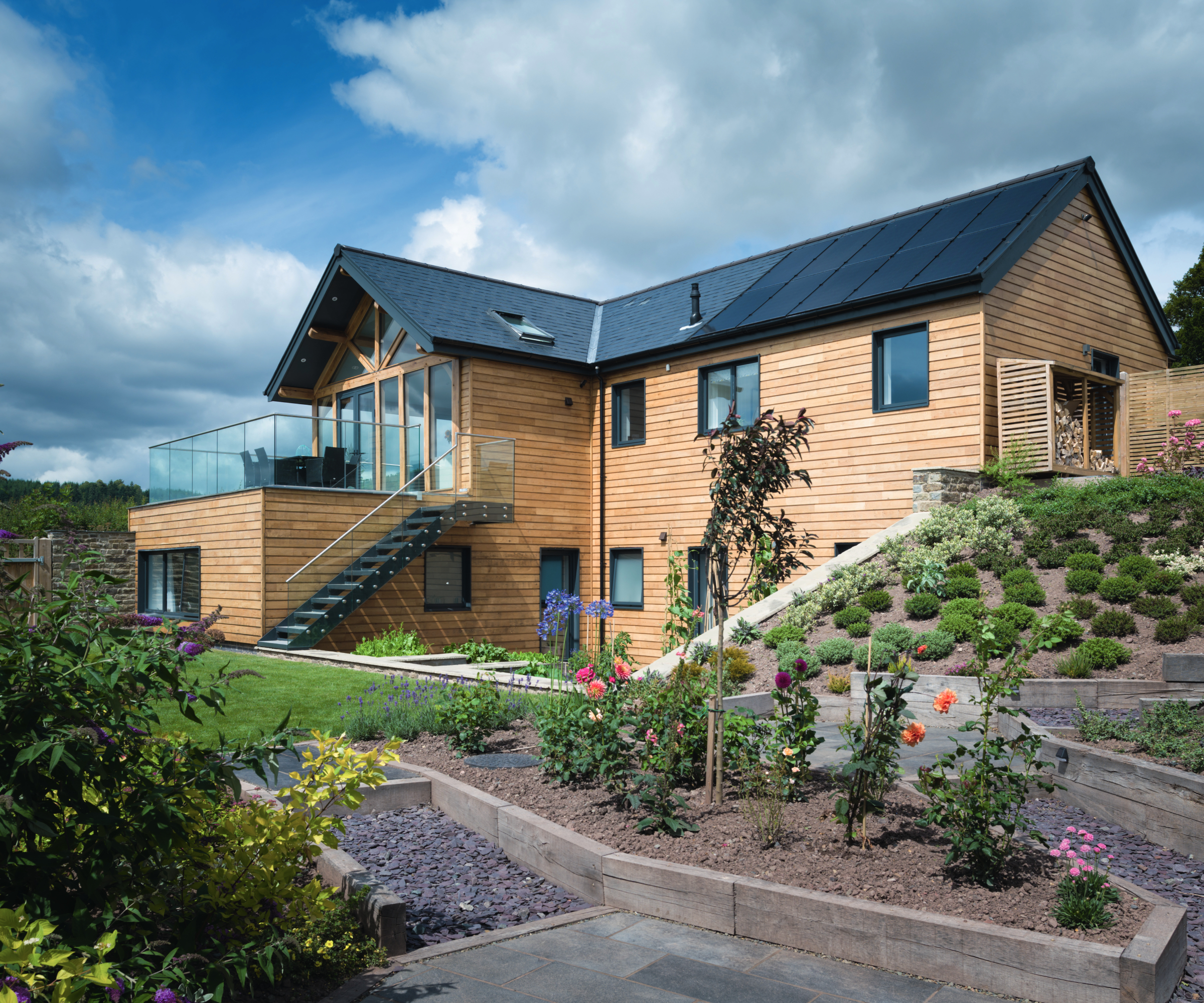 timber clad contemporary home with sloped gardens, terrace and slate roof with solar panels