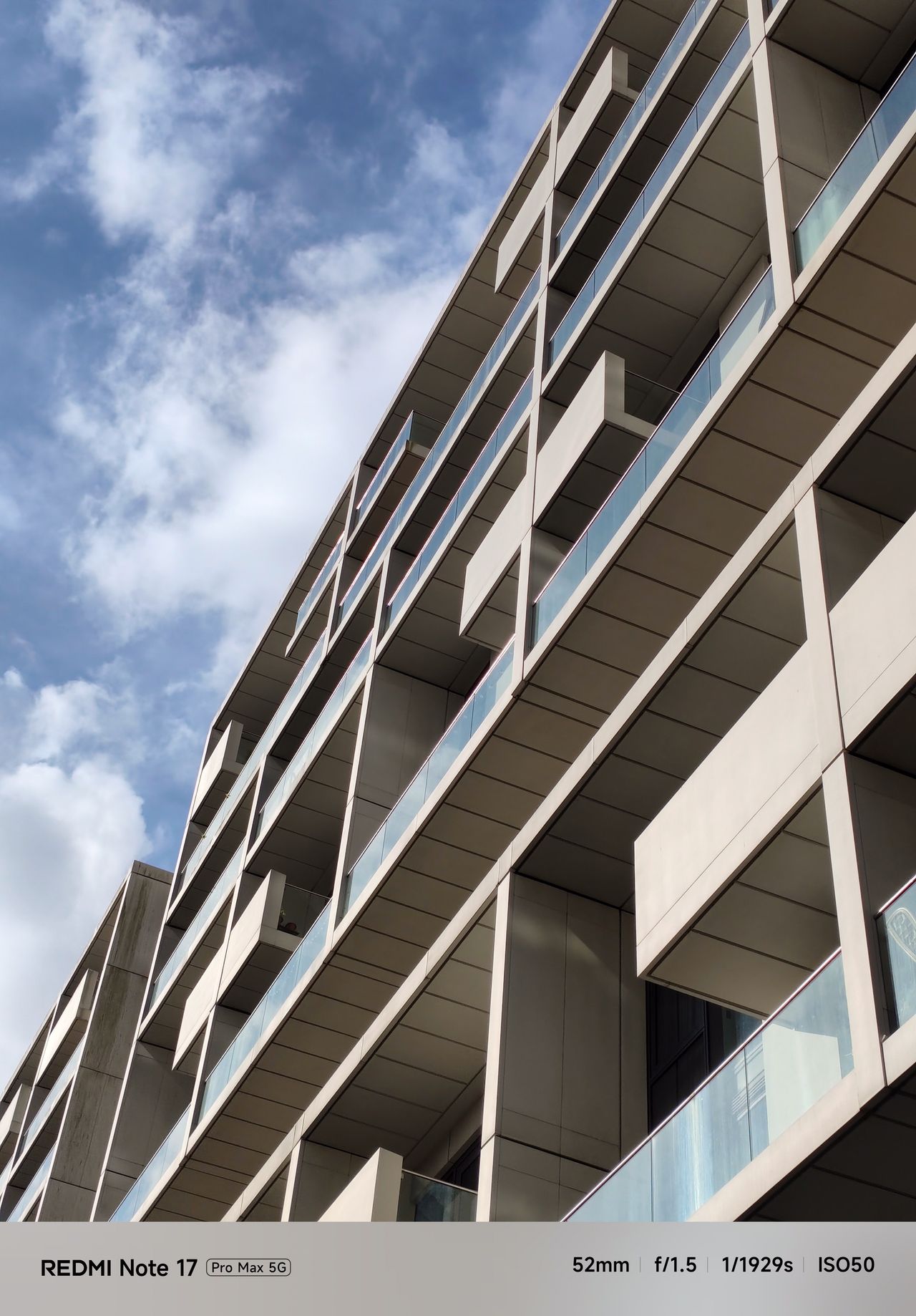 Low-angle 2x zoom photograph of balconies on a modern apartment building against a cloudy blue sky.
