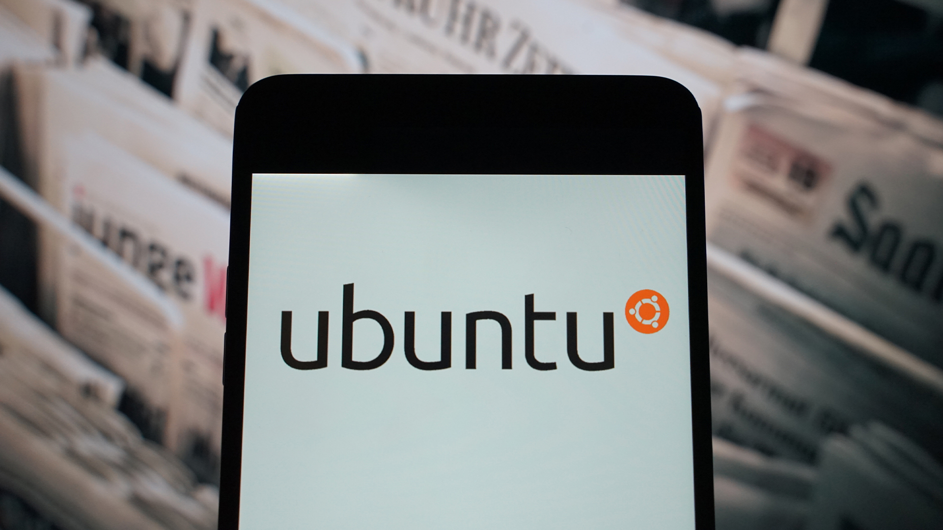 Ubuntu logo and branding pictured on a smartphone screen with newspaper rack with publications in background.