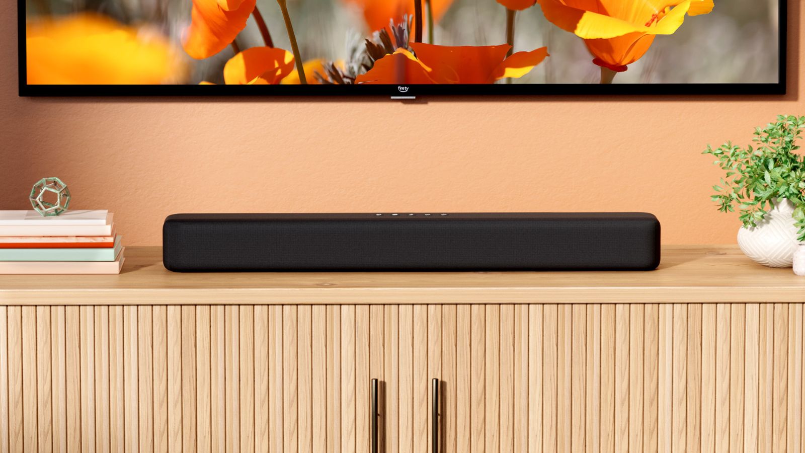Amazon Fire TV Soundbar underneath TV screen showing orange/red flowers, on wooden TV stand next to books and plants, against orange wall.