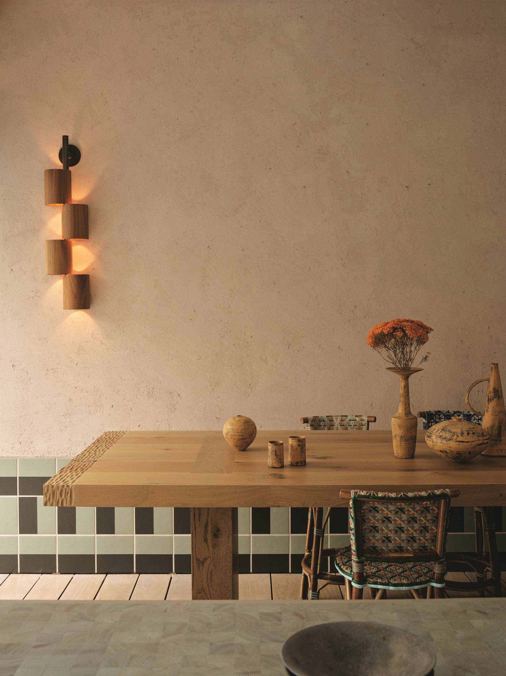 a dining table with textured edge in front of a textured plaster wall with wall sconce and tiled skirting, with woven dining chairs, and styled with vases