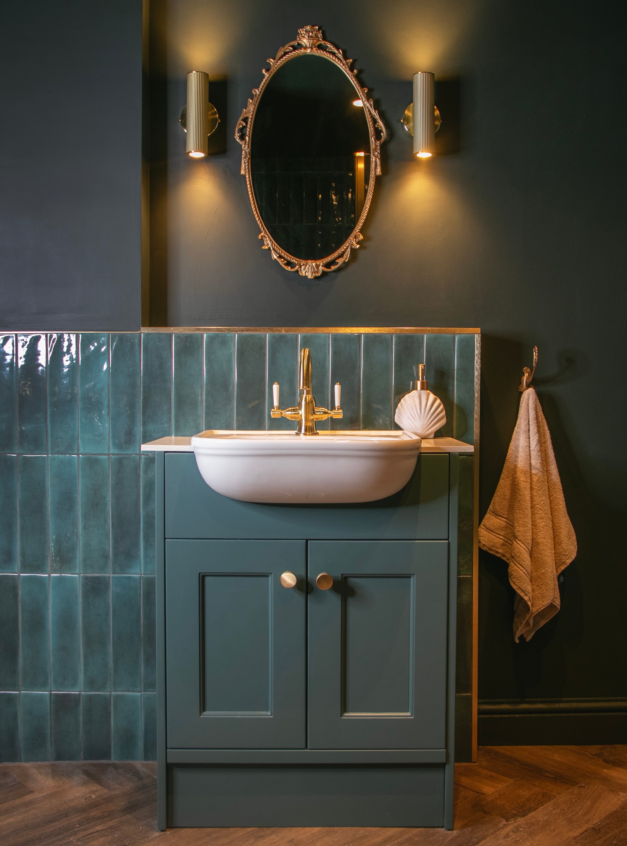 Green color drenched bathroom with vintage style mirror, green wall tiles, two wall sconces on either side of mirror, green vanity cabinetry, white sink, brass taps, and hand towel on loop